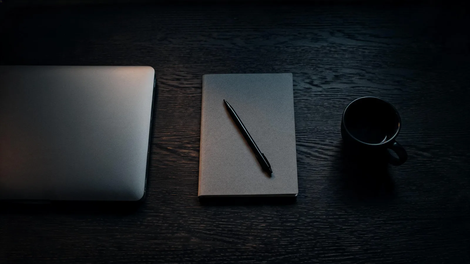 Dunkler Holztisch von oben: links ein zugeklappter silberner Laptop, in der Mitte ein graues Notizbuch mit schwarzem Kugelschreiber, rechts eine dunkle Tasse